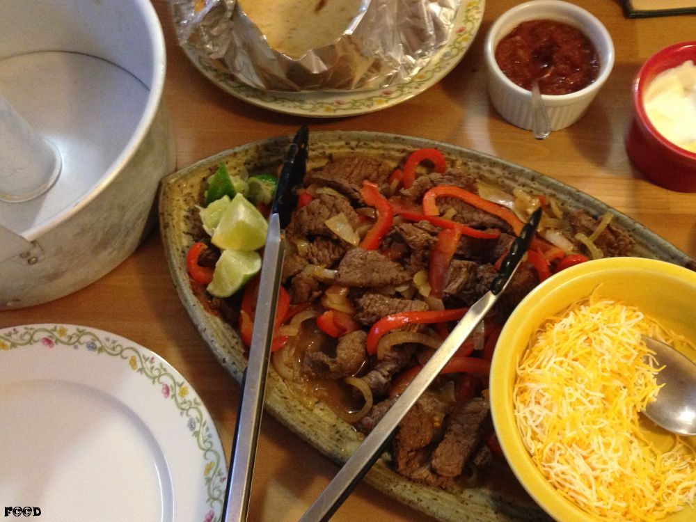 with all the trimmings, steak, pepper, and onions fried in a pan, limes for flavor, heated tortillas, salsa, sour cream, guacamole not shown, shredded cheese, and that all important cake dish to the left