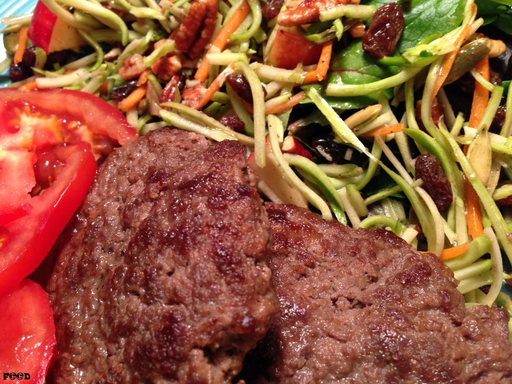 two hamburger patties, a few slices of tomatoes, and a broccoli slaw salad with apples and raisins