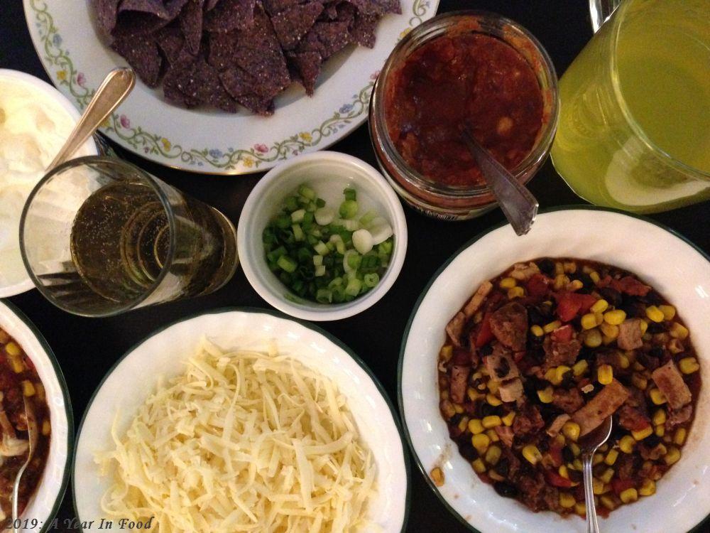 Bowls of chili (black beans, corn, pork slices, hot sausage, tomatoes) surrounded by all the fixingins including green onions, salsa, sour cram, cheese, and chips)