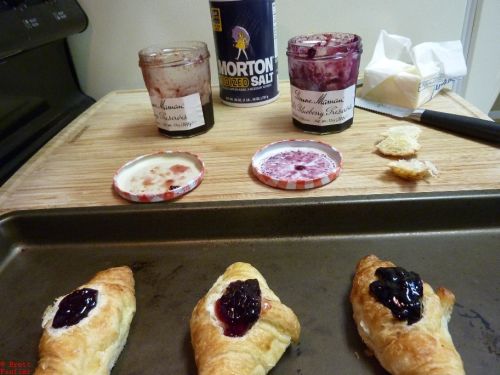 Three croissants in the foreground on a cookie sheet, salt, jelly, butter, sliced tops in the background, ready to go into the oven, brave soldiers that they are, taking one for the team