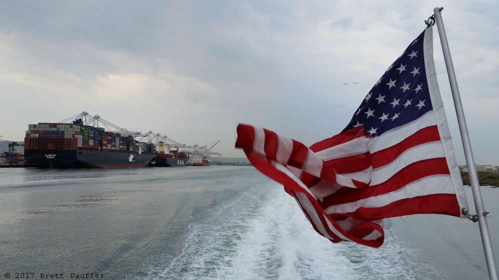 SF Bay on the Ferry from Oakland to SF, looking off the back, container ship to the left, which in this case would be starboard, odd how port was actually on the port side, or SF Harbor, Oakland harbor is what is actually shown, just a little bit, showing the cranes in use, but mostly it is a picture of the American flag