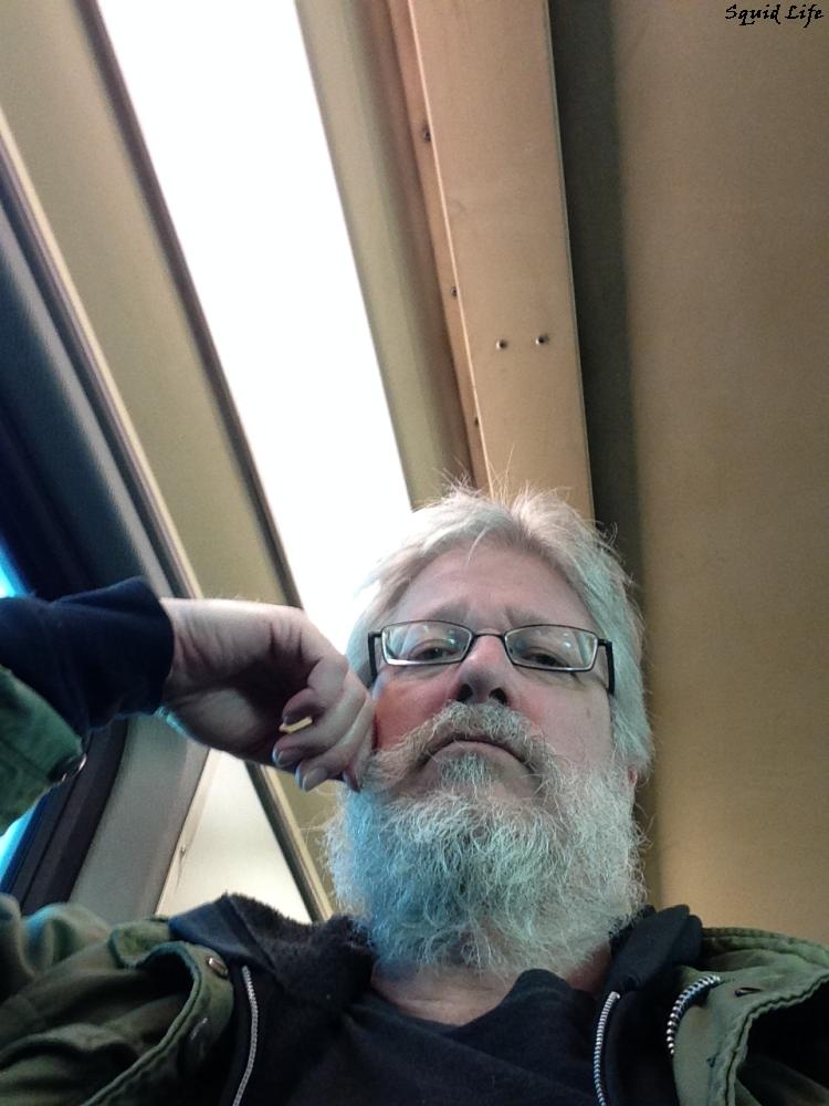 Looking back down, for it is an upward shot, face resting on hand, full beard, mustache, grey hair, lots of grey hair, glasses, black hoodie and army jacket, a typical look of the day