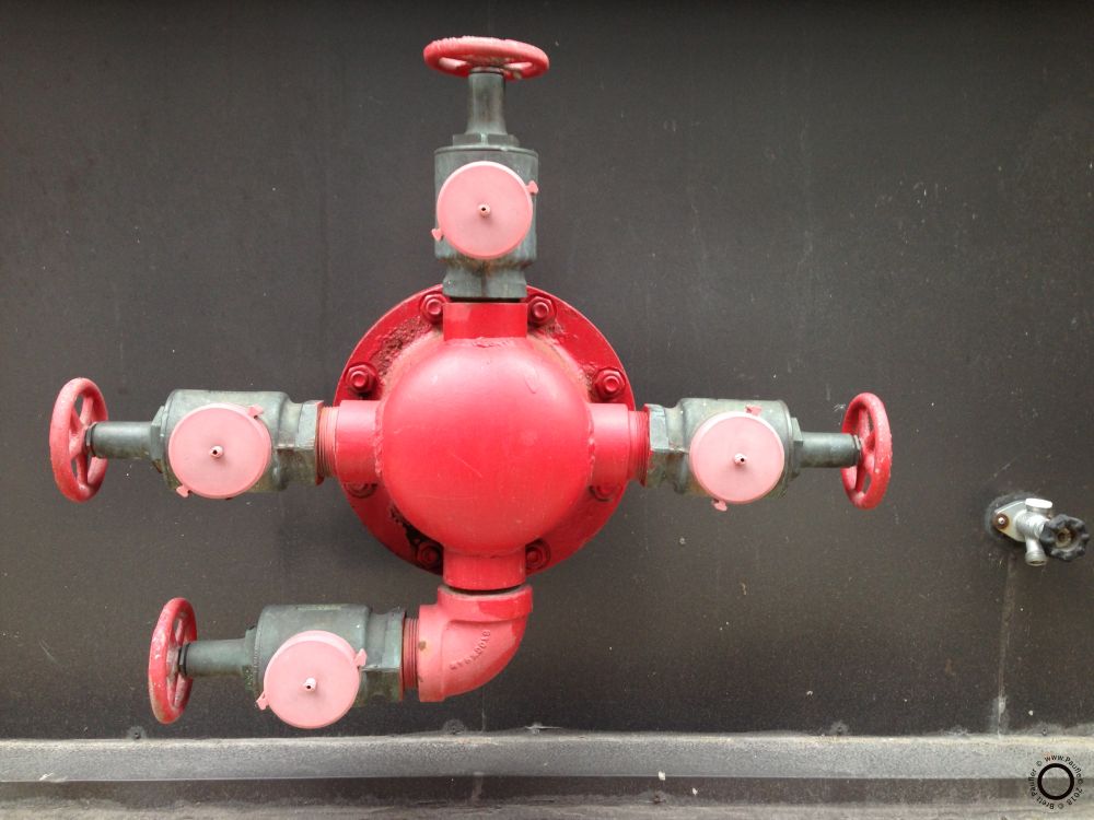 A main water control valve, or so I presume, on the side of a wall, painted all red and pink, so it is pretty like