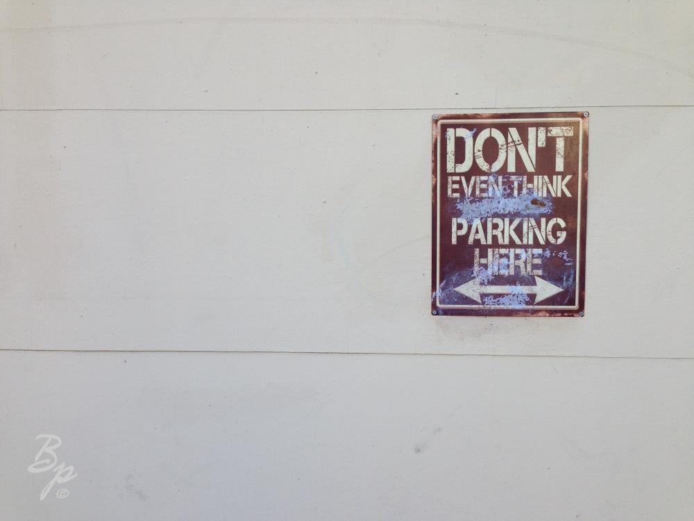 A White Wall with a sign, heavily weathered with paint which reads... Dont Even Think About Parking Here