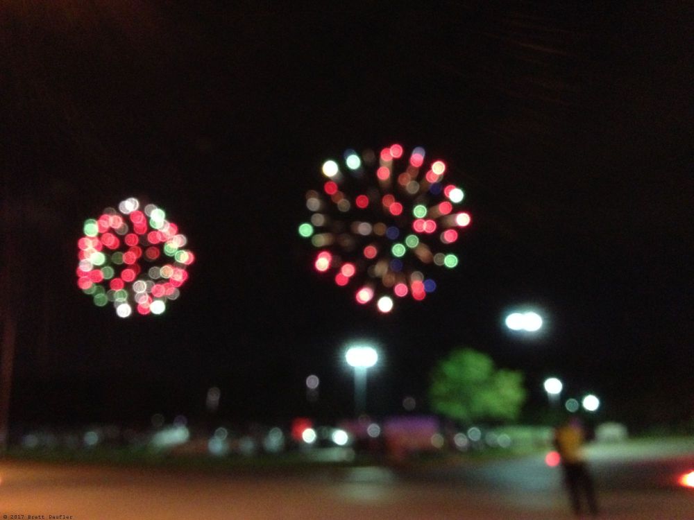 Here we have a blurry view of two fireworks going off, there were three sets, three launch pads of the show we were watching, and here are two, likely a third off screen, or maybe sometimes they only shot off the one or two, anyhow, beyond the road, beyond the police cruiser, it was an amazingly good show