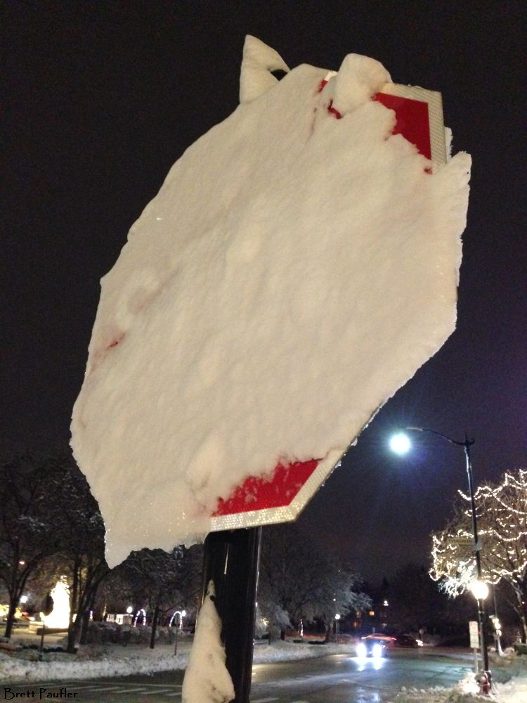 A stop sign covered in snow, but it is not all that bad, as the streets are clear, verified by the car driving by