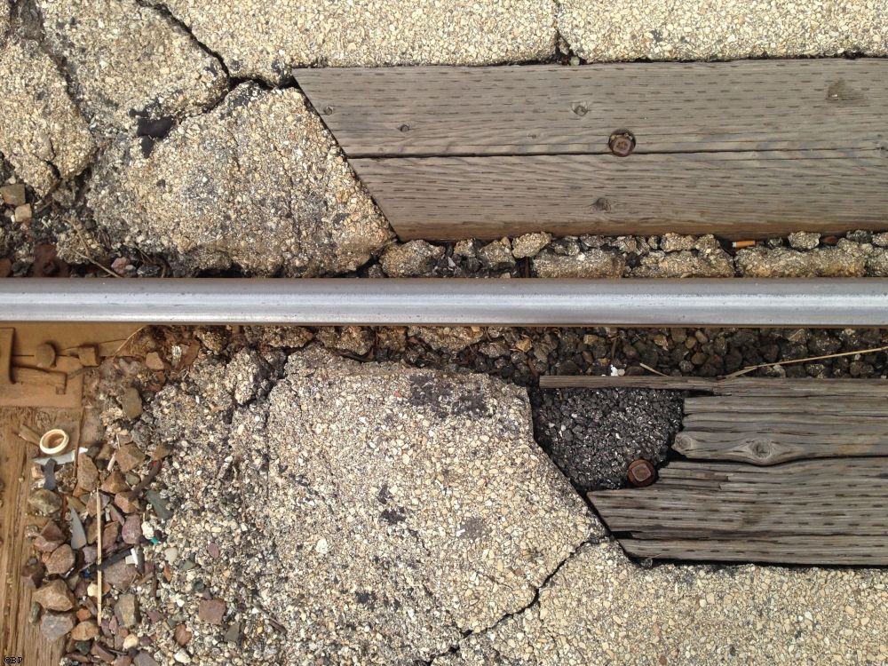 a single rail of a train track cutting through pavement, surrounded by wood nosing, it sort of bothers me that the track is not straight in the image and that the track does not line up with the next image, oh, well