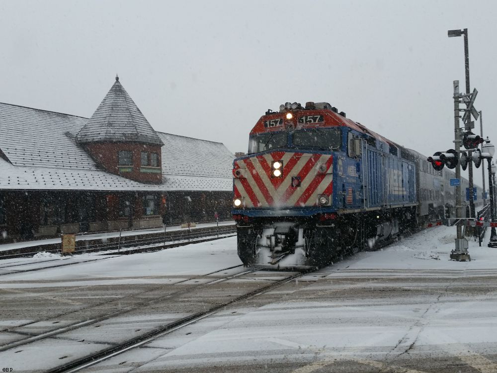 A train coming in with snow in its grill, likely a commuter special, coming in from the city, they had to redo this interesction for the tracks come spring, water got through and expanded under the tracks as it froze, or something, I do not know, but they replaced all of the roadway with prefab pieces of concrete made special for the job to fit between the tracks