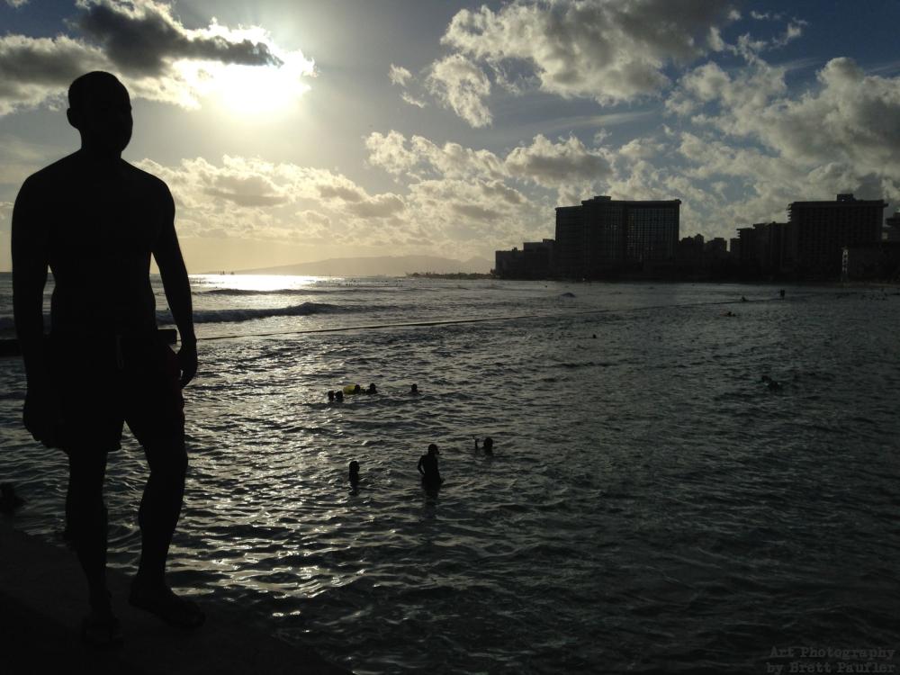 A silhouette of a person standing on pier looking out over ocean, swimmers in the water, buildings in the distance, very much black on muted colors, the shot being taken into the sun