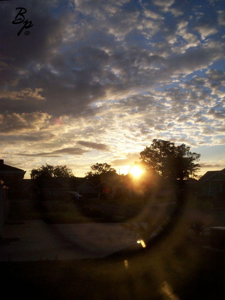 This is an old image from 2009 or so, of a Sunset from the house I used to live in on The Big Island, clouds in the sky, passable sunset, blacked out trees in the foreground, and a nice circular sun flare or light flare from the lens, which really makes the picture, the shot might have been taken through a window, who knows