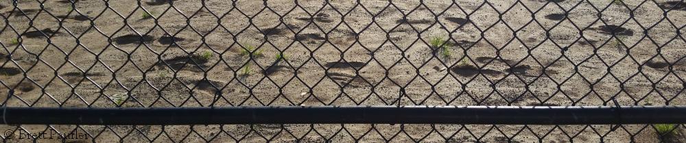 A view through a cyclone fence onto the dirt of a baseball diamond, but it looks more like a beach, this is part of the view from the bench that I favour