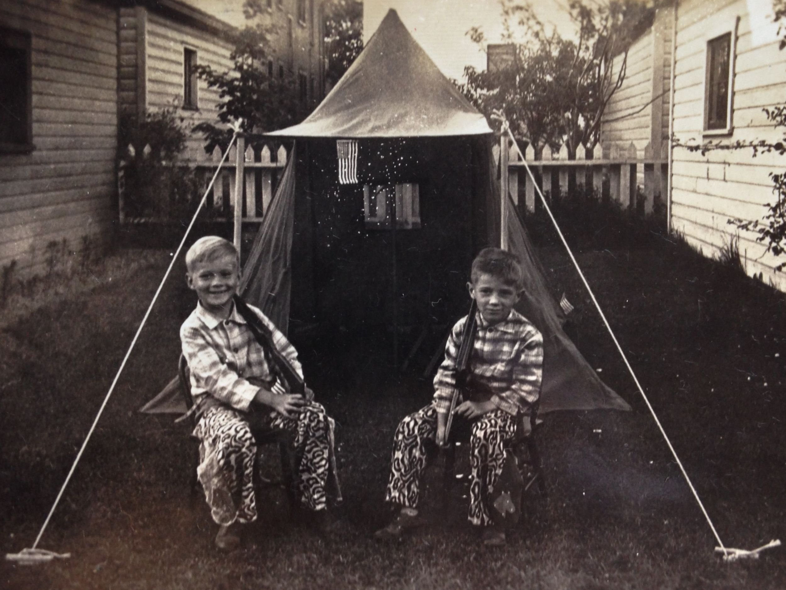 black and white image of two boys playing fort or something like that, World War II era, tent, United States flag, rifles, plaid shirts and weird cowboy leggings, probably cowhide camouflage