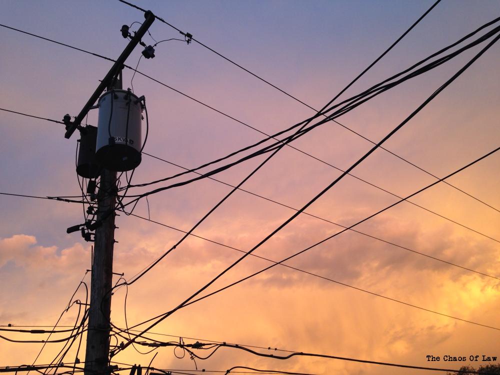 An electric pole (or telephone pole) with all sorts of wires going this way and that against a sunset sky