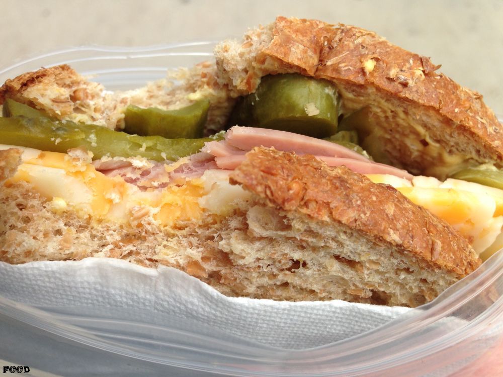 what we have here is a sandwich, standard American, seeded wheat bread, mayonnaise, mustard, colby jack cheese, ham, and lots and lots of pickles, cement sidewalk in the background, as I eat walking down the street