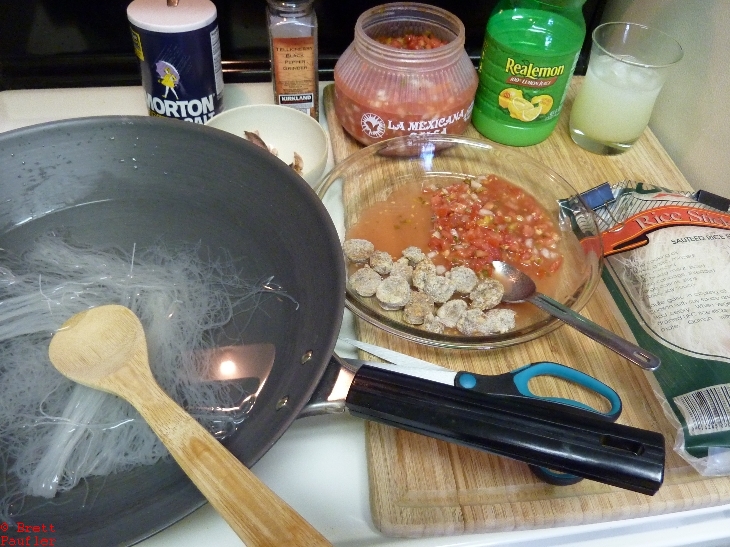 Ingredient shot, those be rice noodles in a wok, I do believe, salt, pepper, gooiing across from the back, salsa, big jug of lemon juice, got the lemonaide going, package of rice noodles, salsa and sausage in a pice dish, scissors up from, the intent to show what I needed in one shot, this is actually a pretty good shot from that perspective