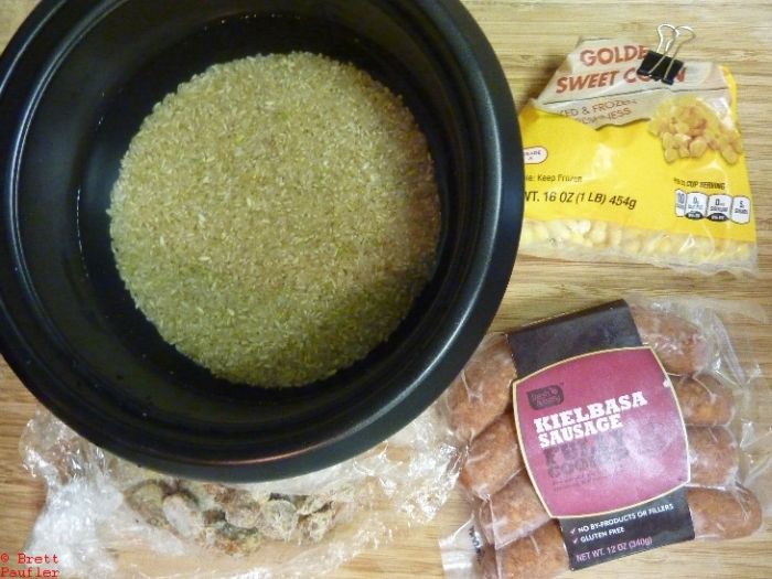 Ingredient shot, rice and water in cooker, corn to side, with sausage and bits of frozen sausage below