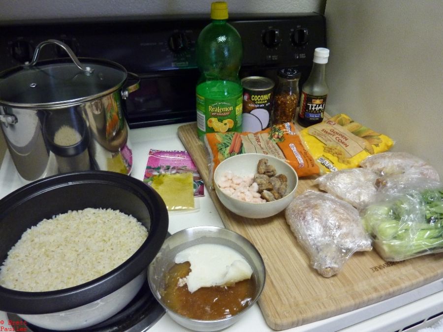 The ingredient shot, rice, meat juice of some sort, lemon juice in a bottle, coconut milk, red peppers, thai fish sauce, frozen peas, frozen corn, bits of sausage in a bowl with shrimp, frozen bock choy, probably three different other types of frozen meat, got no idea what the big bowl is for at this point, probably to mix it all up in after the rice is done