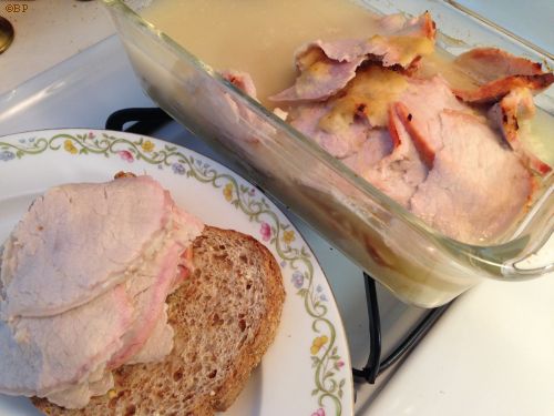 A slice of bread with a mound of sliced pork on it, to the side, a glass dish of pork marinating in horseradish and pickle juice, slightly brown on top