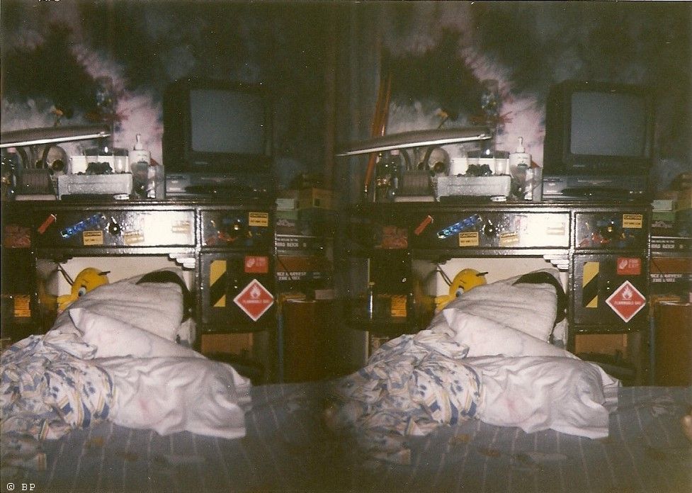 Image of a bed, circa late 20th century, I know not how typical or not, it is a standard twin mattress set directly on the floor with cartoon sheets, tie dye pillow case, tweety bird stuffed bird, and who knows what else, a sticker covered childs desk, all of this was new to me in my twenties or so, tie dye on the wall, tv with recorder, maybe a sega game somewhere, Third Reich stacked with other board games to the side, desk light, skull with newly painted red eyes, I think I did that, and a bunch of miscellanous crap, I doubt I own a single thing pictured in the image anymore