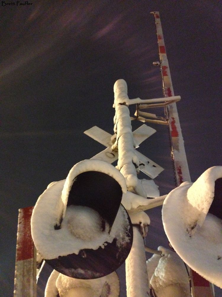 Upright Railroad crossing signal, covered in snow, the bar that comes down in the upright position, highlighting the swirling wisp of clouds far above