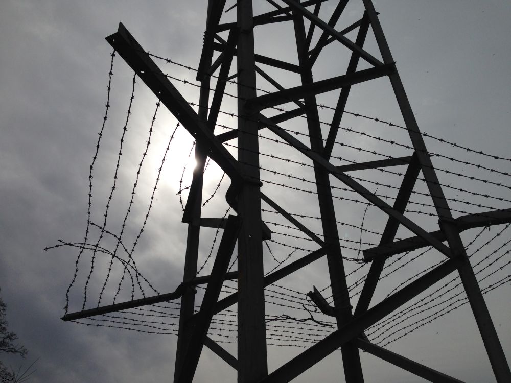 A power line tower, once again, the lower middle half, highlighting the barbed wire fence that prevents climbers, which makes it look more prison-ish, sun in the background on an overcast day