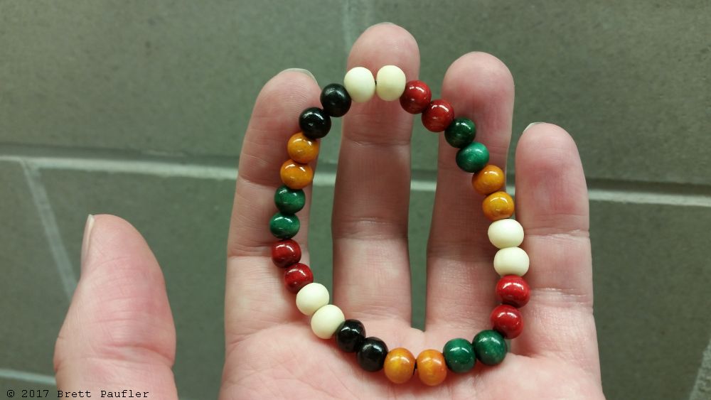 A wooden bracelet, held in my hand, the backdrop being a cinder block wall, nearby where I acquired said item of honour