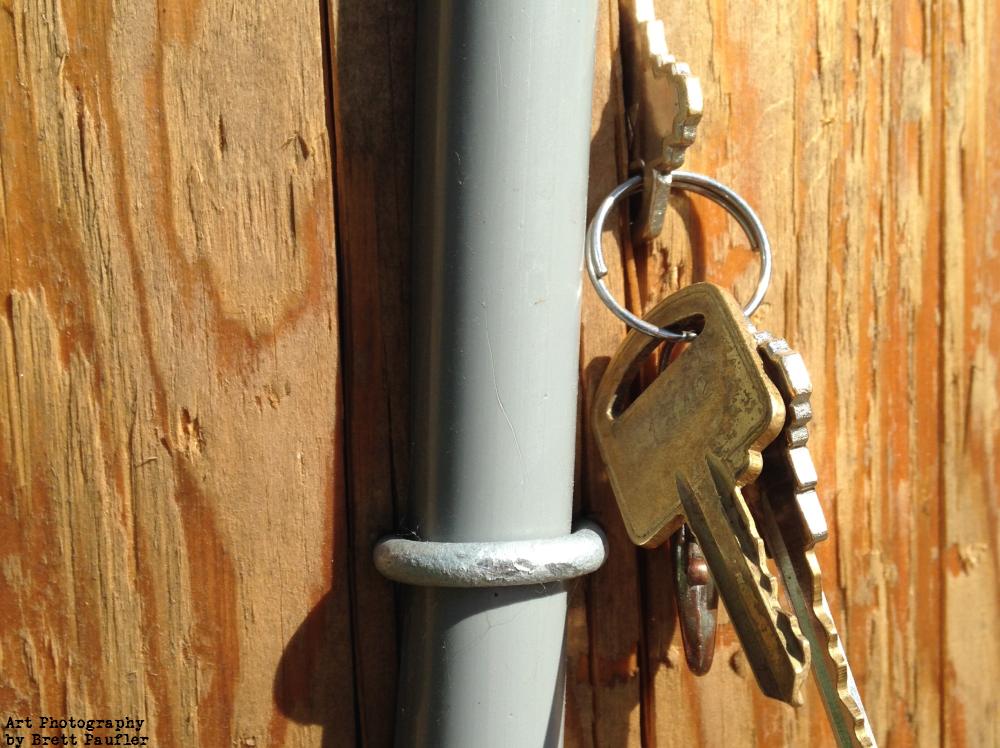 We have a wood background with a pipe going up the center and keys to the right, it is a close up of an electric pole and keys, presumably found, were wedged into the wood for the owner to find