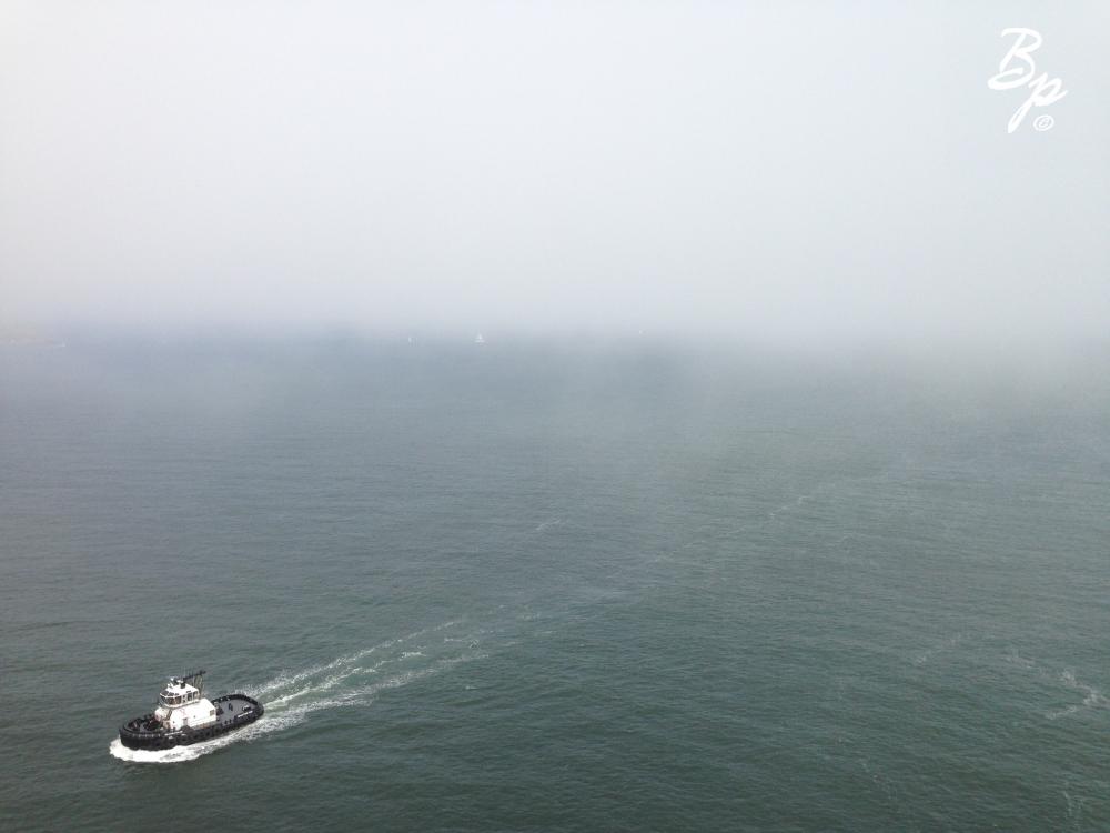 Taken from the Golden Gate Bridge looking down and out, a Tug Boat motors off the bottom left side of the screen, while the background is shrouded in fog