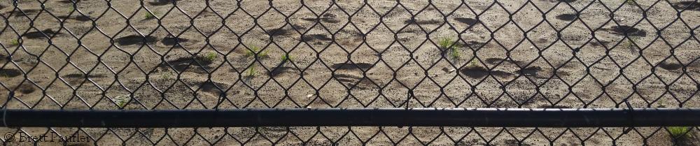 A view through a cyclone fence onto the dirt of a baseball diamond, but it looks more like a beach, this is part of the view from the bench that I favour