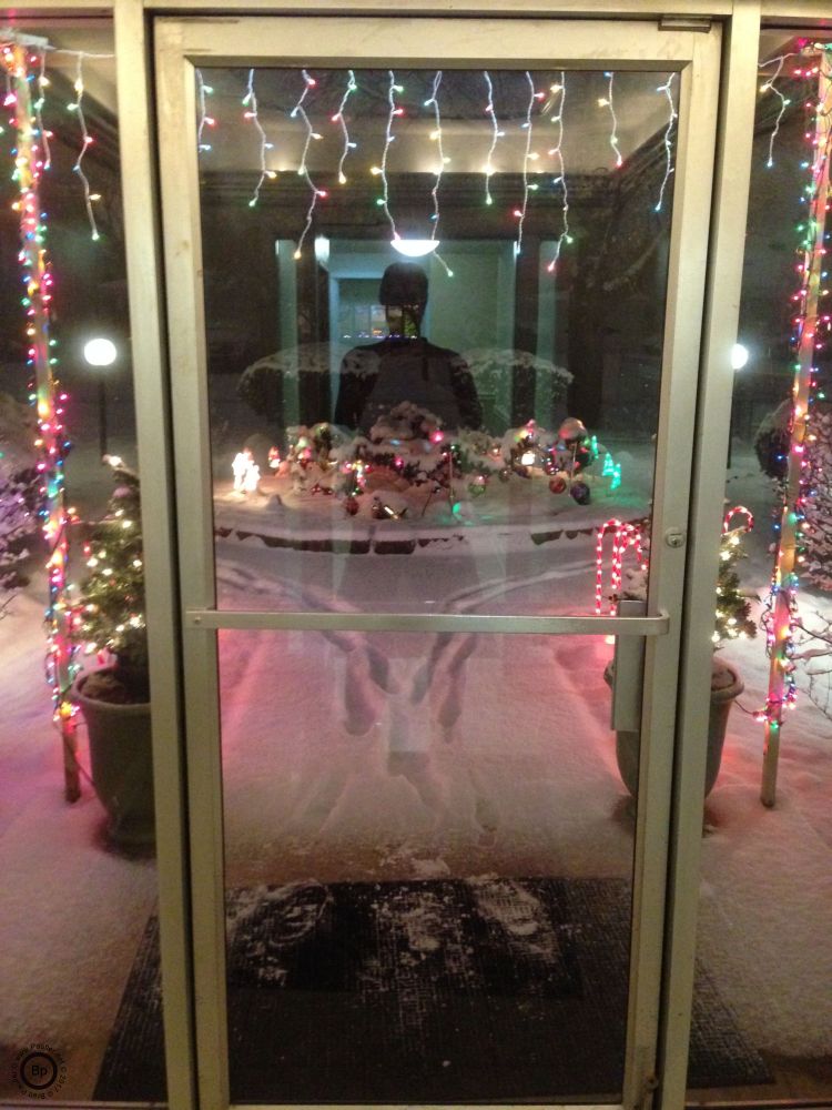 Front door of an apartment complex shown from the inside out, me in reflection, holiday lights lining the opening, snow all about, its a nice pleasing picture