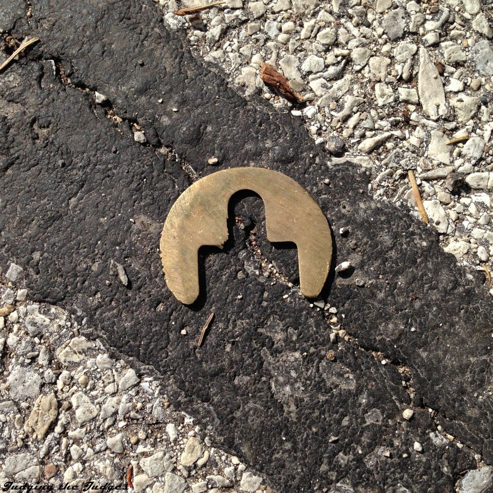 We have a metal plate, brass, likely from a door lock, on a black line of tar, in the middle of the road, well, it is a close up, so who knows where on the grey aging asphalt it is, probably kicked to the side, anyhow, it looks sort of mannish